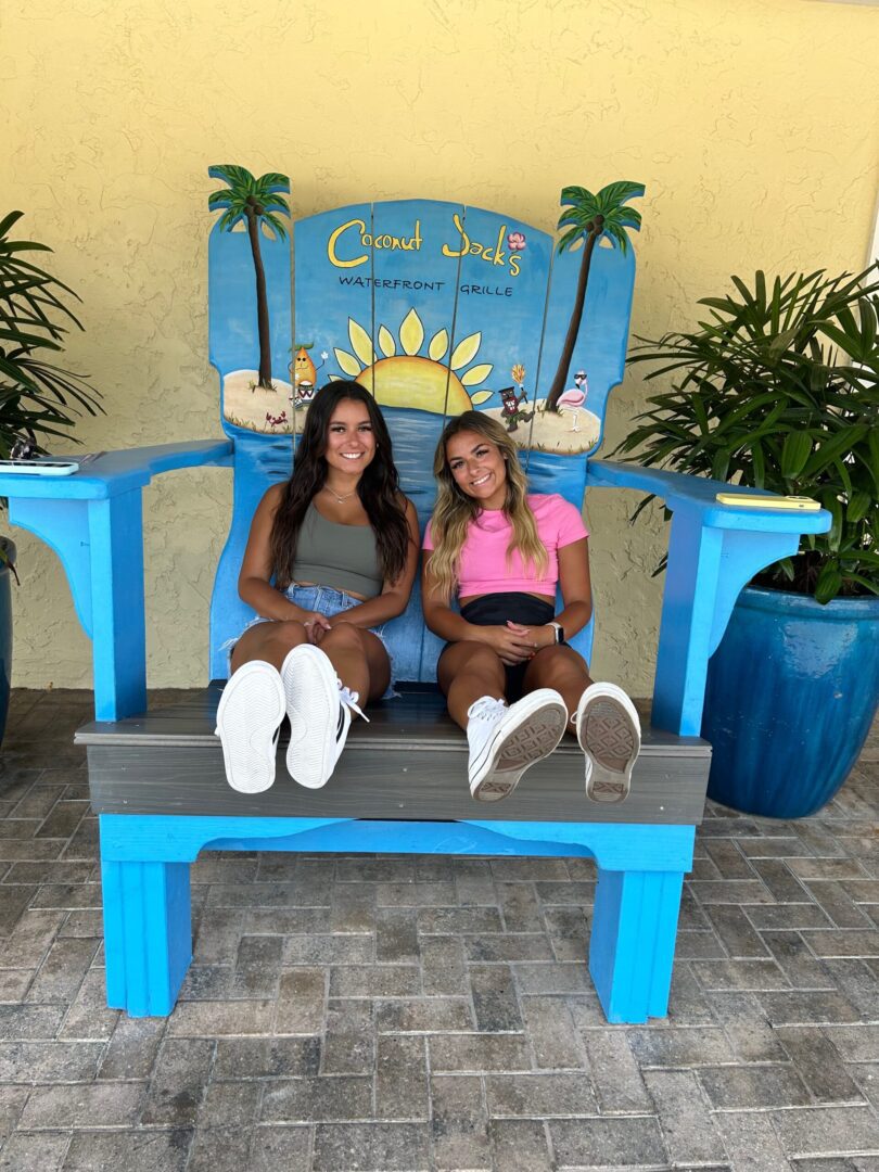 Two women sitting on a giant chair in front of palm trees.